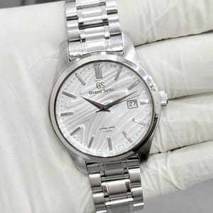 May include: A silver-tone Grand Seiko watch with a white dial featuring a textured, wavy pattern. The watch has a stainless steel link bracelet, silver-tone hour markers, a date window at the three o'clock position, and text reading Grand Seiko, Spring Drive, and 5 Days.
