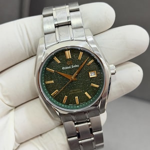 Op de afbeelding: Een Grand Seiko automatisch horloge met een getextureerde donkergroene wijzerplaat, goudkleurige wijzers en uurmarkeringen, en een roestvrijstalen schakelarmband. De wijzerplaat bevat het Grand Seiko logo, een datumvenster en tekst.