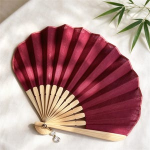 May include: A folding hand fan featuring a deep burgundy fabric canopy with a visible woven texture. The fan is constructed with light-colored bamboo ribs and includes a small, clear, faceted crystal bead charm attached to the base pivot point.