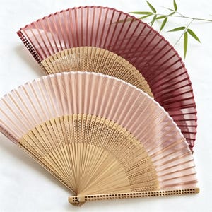 May include: Two folding hand fans with light-colored wooden frames featuring intricate laser-cut geometric patterns. One fan has a deep burgundy fabric canopy, while the other features a soft, pale pink fabric canopy, both displaying vertical pleats.