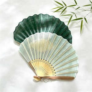May include: Two folding hand fans with a semi-circular shape. One fan is a deep forest green, while the front fan is a pale mint green with a light-colored wooden frame featuring cut-out flower details at the base and a small dangling crystal charm.