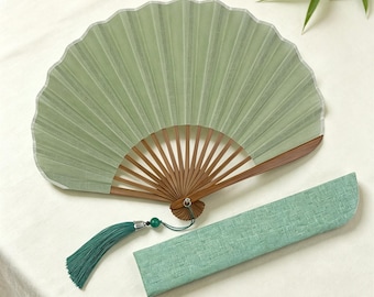 May include: A sage green folding hand fan with a wooden frame and a matching fabric sleeve. The fan features a long, dark teal tassel attached to the base with a small green bead and silver hardware. The fan is fully opened to display the pleated fabric.