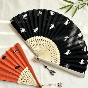 May include: Two folding hand fans with light-colored wooden ribs and decorative tassels. One fan is black with white cat silhouettes and checkered patterns, while the other is orange with black cat silhouettes. Both feature intricate cutouts.