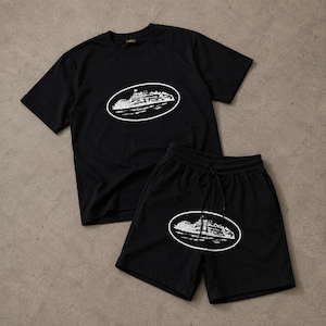 Könnte beinhalten: Ein Set aus einem schwarzen T-Shirt mit kurzen Ärmeln und einer passenden schwarzen Shorts. Beide Teile zeigen einen weißen, ovalen Grafikdruck eines Kreuzfahrtschiffes auf dem Wasser. Die Shorts haben einen elastischen Bund mit Kordelzug.