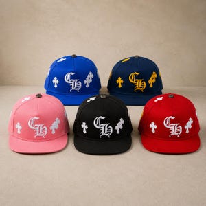 Könnte beinhalten: Fünf Baseballkappen in einem Halbkreis angeordnet, in den Farben Blau, Marineblau, Pink, Schwarz und Rot. Jede Kappe zeigt ein markantes, weiß oder gelb gesticktes CH-Logo auf der Vorderseite, flankiert von Kreuzmotiven und einem Metallknopf.