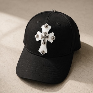 Könnte beinhalten: Eine schwarze Baseballkappe mit einem weißen Leder-Kreuzaufnäher auf der Vorderseite. Das Kreuz ist mit fünf kleinen, runden, silberfarbenen Metallnieten verziert, und eine weitere passende Metallniete befindet sich oben auf der Kappe.
