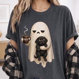 May include: A charcoal gray t-shirt featuring a graphic of a beige ghost with a bored expression holding a brown mug with the text MEH and a steaming liquid, while also cradling a small black puppy in its arm. The ghost has a simple, rounded shape.