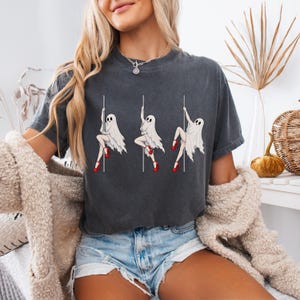 May include: A charcoal gray short-sleeve t-shirt featuring a graphic print of three white ghosts wearing red high heels, each posed on a vertical pole. The ghosts are depicted in different positions while holding onto the poles in a repeating pattern.