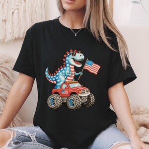 May include: A black t-shirt featuring a graphic of a blue dinosaur with white stars and red stripes, wearing sunglasses and holding an American flag. The dinosaur is standing on a red monster truck with a small American flag decal on the side.