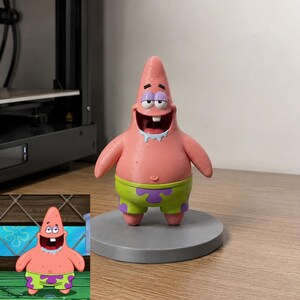 May include: A three-dimensional figurine of a pink starfish character with a wide, open-mouthed smile, a single visible tooth, and light blue drool. The character wears lime green shorts with purple flower patterns and stands on a circular gray base.