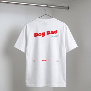 May include: A white short-sleeved t-shirt featuring the red text Dog Dad and green text Essentials! on the upper back. The lower back includes the red text DAISY and smaller text including Dog Lovers Club and Est. 2020.