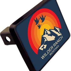 May include: A black and blue hitch cover with a retro sunset design featuring mountains and flying birds. The text "Wilder Ranch Montana" is printed on the cover.