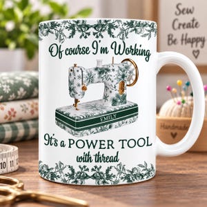 Könnte beinhalten: Ein weißer Keramikbecher mit einer Illustration einer Nähmaschine in grün-weißem Blumenmuster. Der Text lautet: Of course I'm Working, It's a POWER TOOL with thread, und der Name EMILY ist auf dem Sockel der Nähmaschine aufgedruckt.