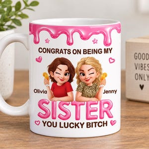 Op de afbeelding: Een witte keramische mok met een roze druppelontwerp aan de bovenkant. De voorzijde toont twee stripfiguren met oranje drankjes, met de tekst: CONGRATS ON BEING MY SISTER YOU LUCKY BITCH. De namen Olivia en Jenny staan bij de personages gedrukt.