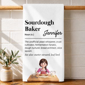May include: A white waffle-textured kitchen towel featuring the black printed text Sourdough Baker Noun (n.) Jennifer, followed by a definition and a cartoon illustration of a person rolling dough on a wooden board with a rolling pin.