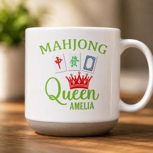 Op de afbeelding: Een witte keramische mok met een grijze voet, voorzien van de groene tekst MAHJONG, een rode kroonafbeelding, de groene tekst Queen en de groene naam AMELIA. Boven de kroon staan drie mahjongtegels met rode, groene en blauwe symbolen op een witte achtergrond.