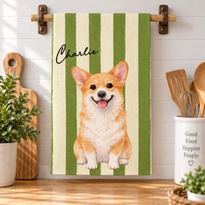 Peut inclure: Un torchon de cuisine à texture gaufrée orné de rayures verticales vertes et crème. Le centre présente une illustration détaillée d'un chien Corgi beige et blanc la langue sortie. Le prénom Charlie est inscrit en lettres cursives noires.
