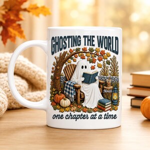 May include: A white ceramic mug featuring a graphic of a ghost sitting in a wooden chair reading a book under a tree. The text reads GHOSTING THE WORLD and one chapter at a time. Pumpkins, a lantern, and a stack of books surround the ghost.
