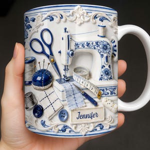 May include: A white ceramic mug featuring a blue and white embossed design of sewing tools including a sewing machine, scissors, a pincushion, a thimble, buttons, a measuring tape, and a fabric swatch with the name Jennifer printed on a label.