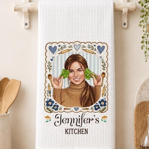Peut inclure: Un torchon de cuisine blanc à texture gaufrée orné d'une illustration centrale représentant une personne tenant de la laitue, encadrée de cœurs bleus, de fleurs et de cuillères en bois. Le texte "Jennifer's KITCHEN" est inscrit en dessous.