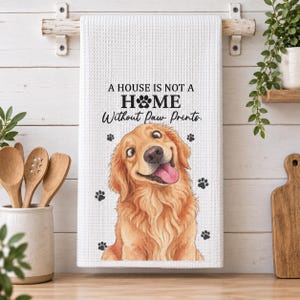 Könnte beinhalten: Ein weißes Küchentuch mit Waffelstruktur, das eine Illustration eines Golden Retrievers mit herausgestreckter Zunge und schwarzen Pfotenabdrücken zeigt. Der Text lautet A HOUSE IS NOT A HOME Without Paw Prints, wobei ein Pfotenabdruck das O ersetzt.