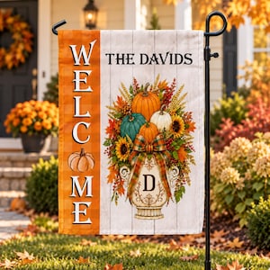 Könnte beinhalten: Eine rechteckige Gartenflagge mit einem vertikalen orangefarbenen Karostreifen links mit dem Wort WELCOME und einer Kürbisgrafik. Rechts stehen THE DAVIDS, eine dekorative Urne mit einem D und ein herbstliches Kürbisarrangement.