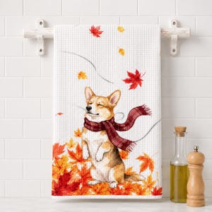 Op de afbeelding: Een witte theedoek met wafelstructuur voorzien van een aquarelillustratie van een bruin-witte Corgi-hond met een rood en donkerrode sjaal, staand te midden van een hoop levendige oranje, rode en gele herfstbladeren van de esdoorn.