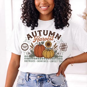 Può includere: Una t-shirt bianca con una stampa grafica che riporta il testo "AUTUMN Harvest", "ORCHARD FRESH", "HAND PICKED DAILY" e "HAYRIDES • BONFIRES • SWEET TEA". Il design include illustrazioni di due zucche, un girasole e spighe di grano.