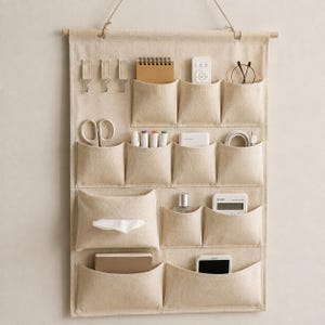Può includere: Un organizer da parete in tessuto beige con tasche multiple e tre ganci in metallo. L'organizer contiene vari oggetti, tra cui un taccuino, un telecomando, occhiali, forbici, pennarelli, una scatola di fazzoletti, una calcolatrice e uno smartphone.
