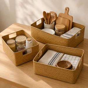 May include: Three rectangular woven storage baskets with integrated handles. The baskets contain kitchen items including glass jars with grains, a white ceramic utensil holder with wooden spoons, wooden cutting boards, and folded kitchen towels.