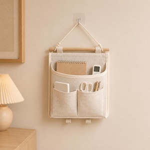 May include: A beige fabric wall hanging organiser with a wooden dowel and a white cord for suspension. It features one large upper pocket containing a notebook and a remote, and two smaller lower pockets holding a white case, scissors, and pens.