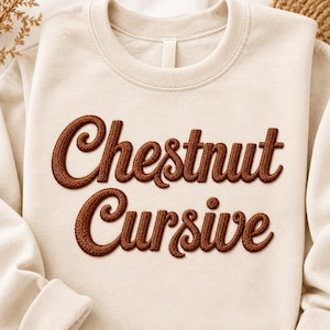 May include: A cream-colored crewneck sweatshirt featuring the words Chestnut Cursive embroidered in a dark brown, raised, textured thread across the chest. The garment has long sleeves, a ribbed neckline, and a soft, solid-colored fabric finish.