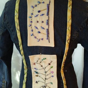 May include: A dark blue denim jacket with decorative embellishments. Two rectangular fabric patches are pinned to the back, adorned with beaded designs. The jacket features yellow trim and beaded accents.