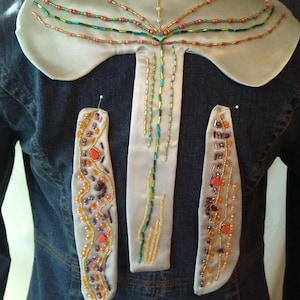 May include: A denim jacket with an elaborate beaded design on the back and sleeves. The design features a white satin collar and three decorative panels, all adorned with colorful beads in shades of red, green, orange, and gold. The beads are arranged in intricate patterns.