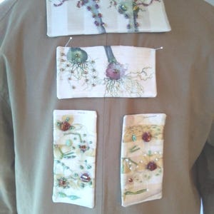 May include: A tan jacket with four decorative fabric patches pinned to the back. The patches are rectangular and feature embroidered designs with beads in shades of green, purple, and red. The jacket is made of a smooth, neutral-toned fabric.
