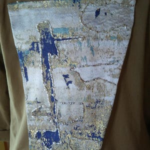 May include: A decorative patch with a blue, gold, and white abstract design. The patch is sewn onto a tan fabric. The design includes beaded accents and a textured appearance, creating a unique visual appeal.