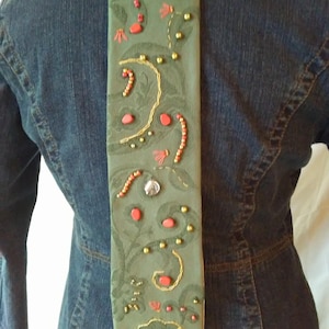 May include: A decorative, olive green tie with floral embroidery, red beads, gold accents, and clear crystal embellishments. The tie is displayed against a denim jacket.