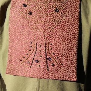 May include: A square, pink textile with a repeating pattern of small, raised circles. The textile is embellished with brown and purple beads, and brown thread, arranged in a tree-like design. The textile is pinned to a tan garment.