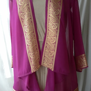 May include: A magenta sheer cardigan with gold trim featuring a floral pattern. The cardigan has a draped front and long sleeves.