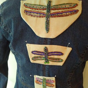 May include: A denim jacket with three dragonfly appliques. Each dragonfly is made of colorful beads and is sewn onto a beige fabric patch. The jacket is a medium wash blue.