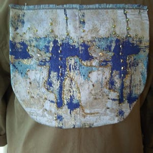 May include: A decorative pocket with a blue, gold, and white abstract design. The pocket is attached to a tan jacket. The design features blue brushstrokes and gold embellishments. The pocket's edges are finished with a blue trim.