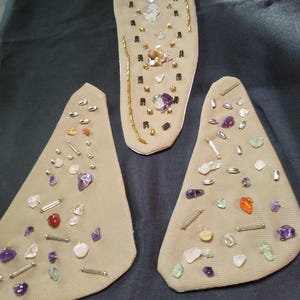 May include: Three beige fabric pieces adorned with various gemstones and metallic accents. The largest piece is oval-shaped, while the other two are triangular. Gemstones include amethyst, quartz, and other colorful stones.