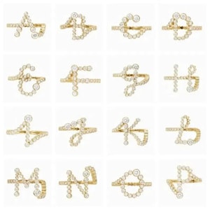 Puede incluir: Una colección de dieciséis anillos de oro, cada uno con una letra diferente del alfabeto de la A a la P. Cada anillo está elaborado con una banda de oro fina y decorado con pequeñas piedras redondas y transparentes que forman la letra.
