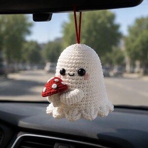 May include: A white crocheted ghost ornament hanging from a red string. The ghost has black bead eyes, a small stitched smile, and pink cheeks. It is holding a small red mushroom with white spots in its arms, featuring a scalloped bottom edge.