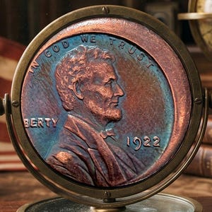 Pode incluir: Um relevo circular em tom de cobre de uma moeda de um centavo Lincoln de 1922 montado numa moldura rotativa de latão. A moeda apresenta o perfil de Abraham Lincoln, o texto IN GOD WE TRUST, a palavra BERTY e o ano 1922 numa base circular.