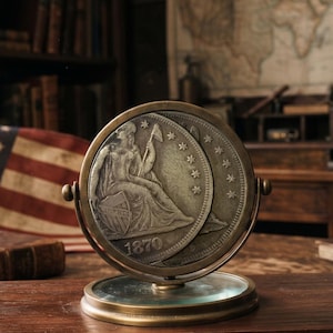 May include: A circular brass display stand holding a silver-toned coin featuring a seated figure holding a pole with a cap, a shield inscribed with LIBERTY, and the year 1870. The coin is positioned vertically within a rotating metal frame.