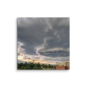 Puede incluir: Un lienzo cuadrado que muestra un cielo dramático lleno de nubes de tormenta grises y pesadas sobre el horizonte. Debajo de las nubes, se observa una línea de árboles verdes y un edificio de varias plantas con fachada de ladrillo al atardecer.
