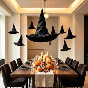 May include: A collection of black pointed witch hats suspended from the ceiling above a long wooden dining table. The table is set with black plates, clear glassware, white pillar candles, orange pumpkins, and autumn leaves on a runner.
