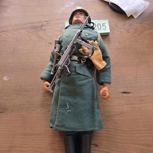 May include: A military action figure wearing a grey helmet, a long grey coat, and black boots. The figure holds a grey submachine gun across its chest and wears tan gloves. A small green rectangular block with the number 205 is visible behind the figure.