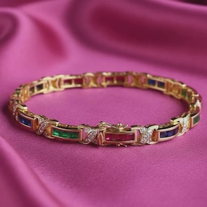 Puede incluir: Una pulsera de eslabones dorados con piedras preciosas rectangulares alternas en rojo, verde y azul. Cada eslabón está separado por un conector en forma de X incrustado con pequeñas piedras claras, y cuenta con un cierre de caja seguro.